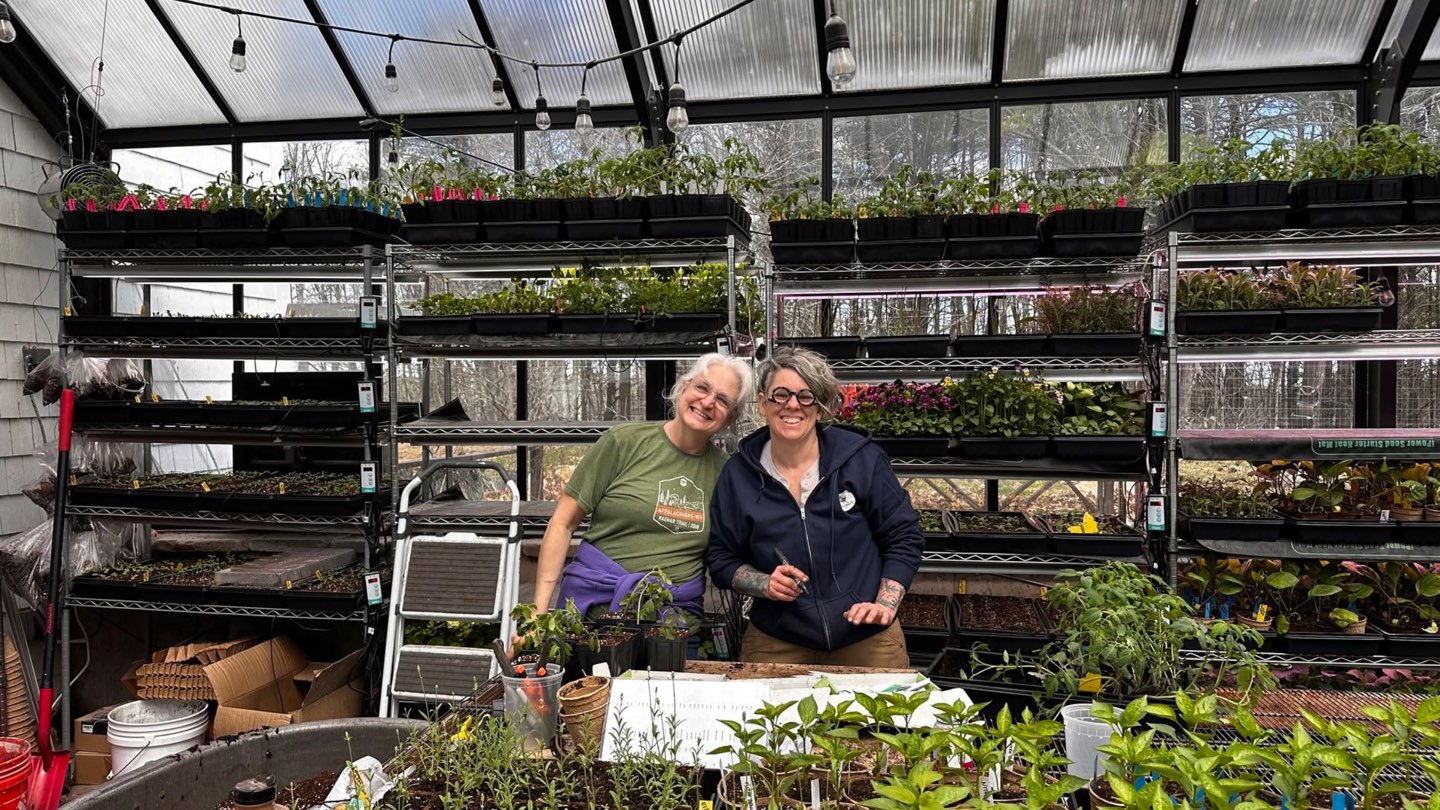 Two people smiling among trays of seedlings in The Laurels Farm greenhouse.